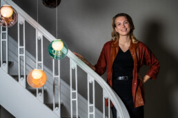 Kvinde smilende på trappe omgivet af farverige glaskuglelamper – moderne og kreativt miljø med karakterfuld arkitektur.