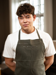 Smilende mandlig kok eller tjener i hvid skjorte og sort forklæde, stående i Gastromés loungeområde med dæmpet belysning.