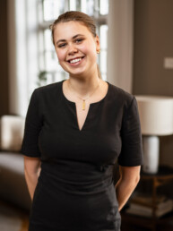 Portræt af smilende kvinde i sort arbejdsuniform i moderne restaurantmiljø, klar til at byde gæster velkommen.