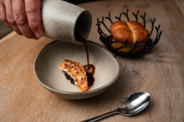 Hånd hælder mørk sauce over delikat fiskeret med sprød skorpe – serveret i keramikskål med friskbagt brioche i baggrunden.