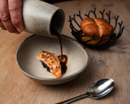 Hånd hælder mørk sauce over delikat fiskeret med sprød skorpe – serveret i keramikskål med friskbagt brioche i baggrunden.