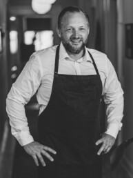 Portræt af smilende kok i hvid skjorte og mørkt forklæde – professionel og indbydende stemning i restaurantens gangareal.