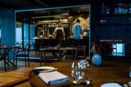 Moderne restaurant med åbent køkken og to kokke i arbejde – elegant indretning med træborde og dæmpet belysning.