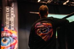 Mand i sort trøje med farverigt øjemotiv, ser mod scenen under et AI-event arrangeret af Klean Group.