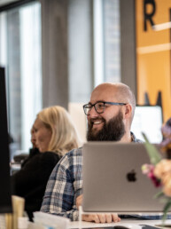 Mand med skæg og briller smiler ved sit skrivebord i et moderne kontormiljø med en åben laptop foran sig