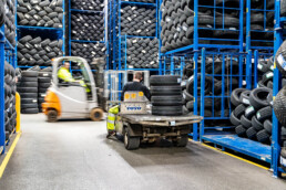 Lagerarbejder transporterer dæk med elbil, mens gaffeltruck opererer i baggrunden. Effektiv dækhåndtering i logistikcenter.