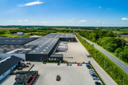 Luftfoto af NDI’s hovedlager og lastbilramper omgivet af grønne marker. Dronebillede af dansk industribyggeri og transporthub.