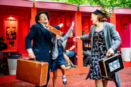 Livlig teatergruppe med kufferter og vintage-tøj underholder festivalgæster ved farverige boder på Smukfest. Foto af Kasper Hornbæk.