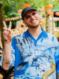 Smilende festivalgæst iført blå mønstret skjorte laver fredstegn ved Smukfest. Glad stemning fanget af Kasper Hornbæk.
